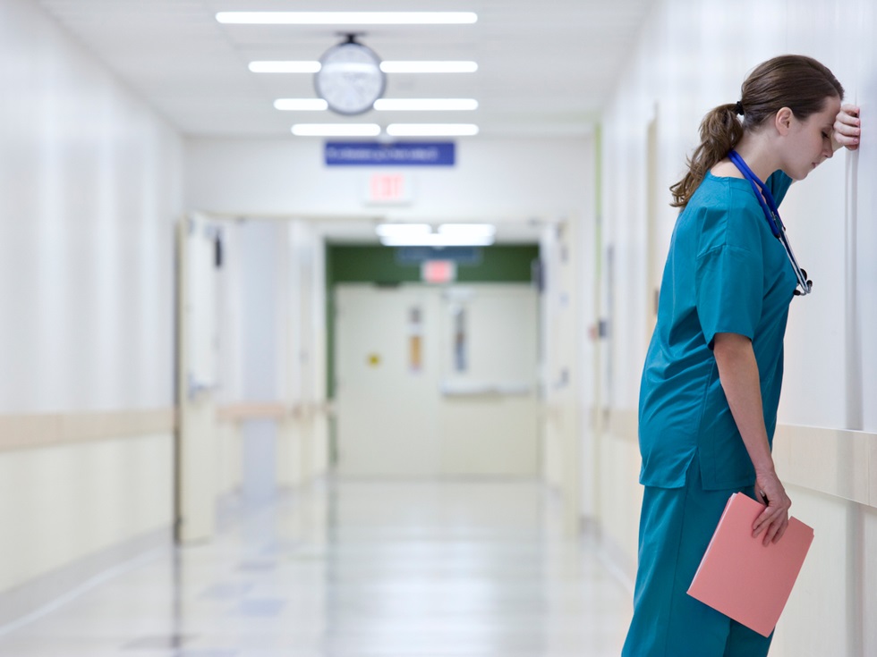 En kvinna med arbetskläder står med huvudet lutat mot väggen i en sjukhuskorridor.