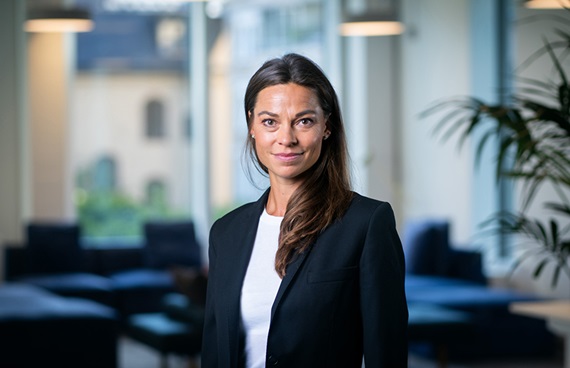 Mörkhårig kvinna i kontorslandskap som tittar in i kameran och ler trevligt.