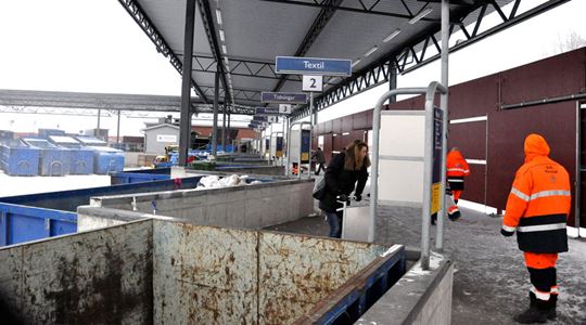 Återvinningsstation med olika containrar och personal i varselkläder.